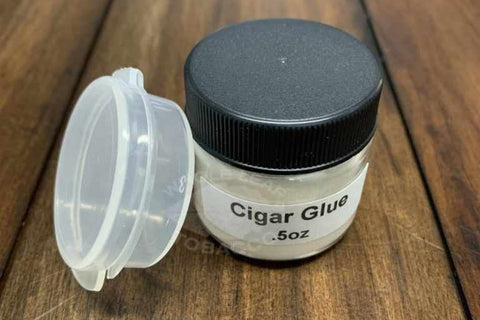 A small, clear glass jar with a black lid labeled "Cigar Glue .5oz" sits next to its translucent cap on a wooden surface.
