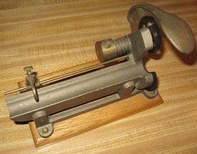 A gray metal cigar tuck cutter with a brass fitting and a curved handle, mounted on a wooden base.