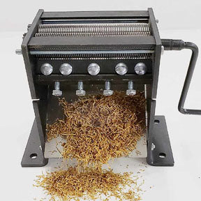 A heavy-duty manual shredder, black with a silver handle, outputs shredded tobacco onto a white surface.